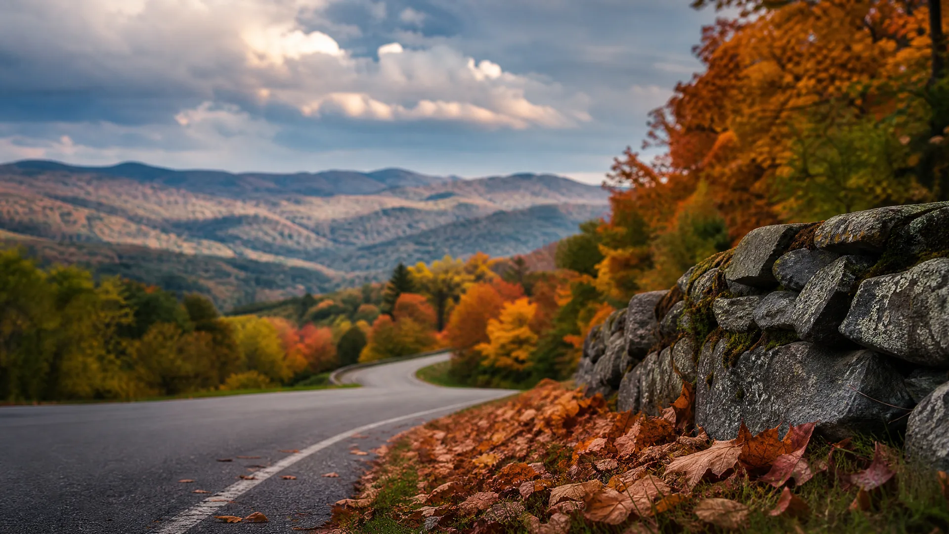 A curving paved road edged by a dry stone wall and fallen leaves, with autumn-colored trees and rolling hills under a cloudy sky.