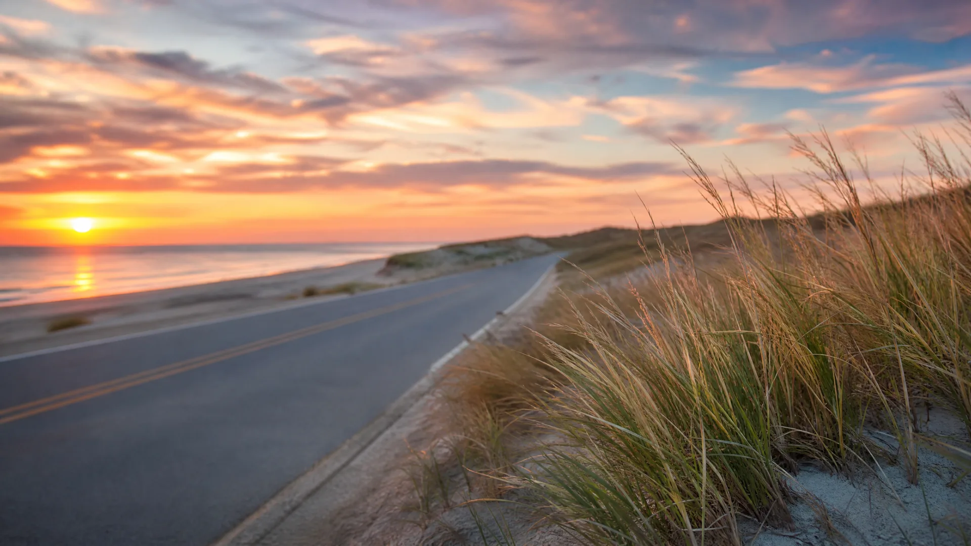 A two-lane road with a yellow center line curves along a coastal dune covered in beach grass, with the ocean and a sunset sky on the left.