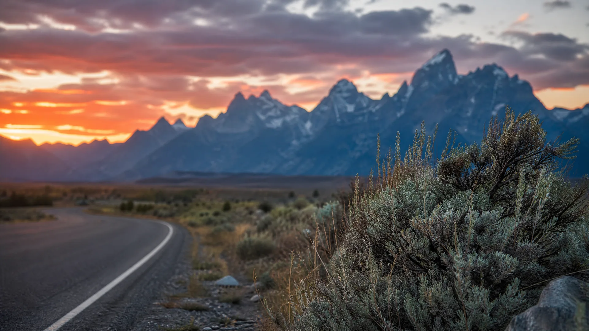 A curving paved road with a white edge line runs past sagebrush toward a jagged, partly snow-covered mountain range under an orange and purple sunset sky.